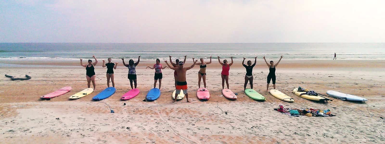 Adult surf class celebrating with arms and shakas raised behind a row of colorful soft-top surfboards at the shoreline