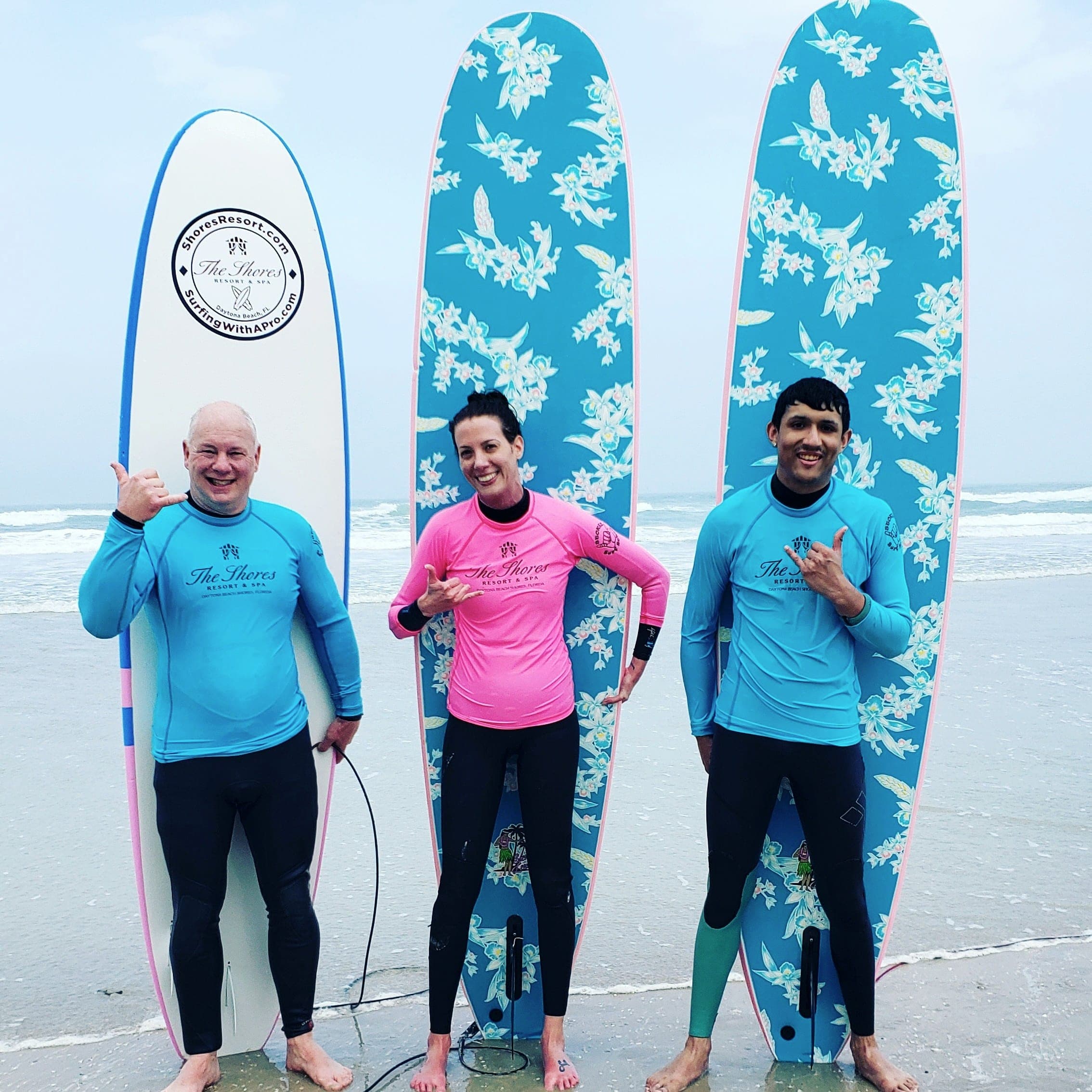 Three adult students with tall soft-top longboards at the shoreline throwing shakas, Daytona Beach