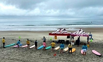 Lesson group lined up with boards at the beach tent, seen from a distance