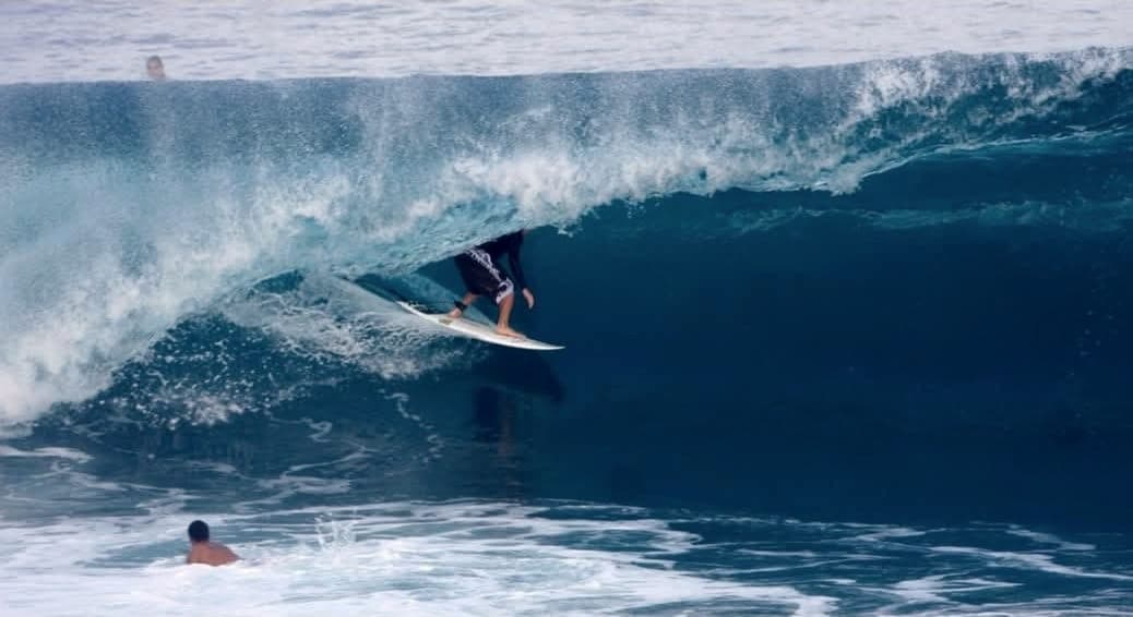 Ryan Ragan riding under the lip of a blue wave, a swimmer visible in the foreground