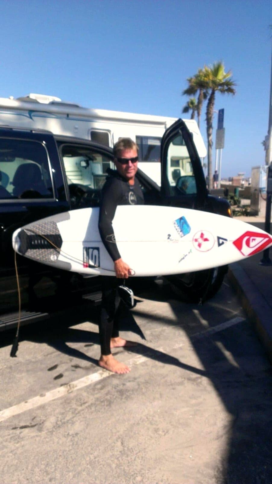 Ryan Ragan in a wetsuit holding his board upright beside a van