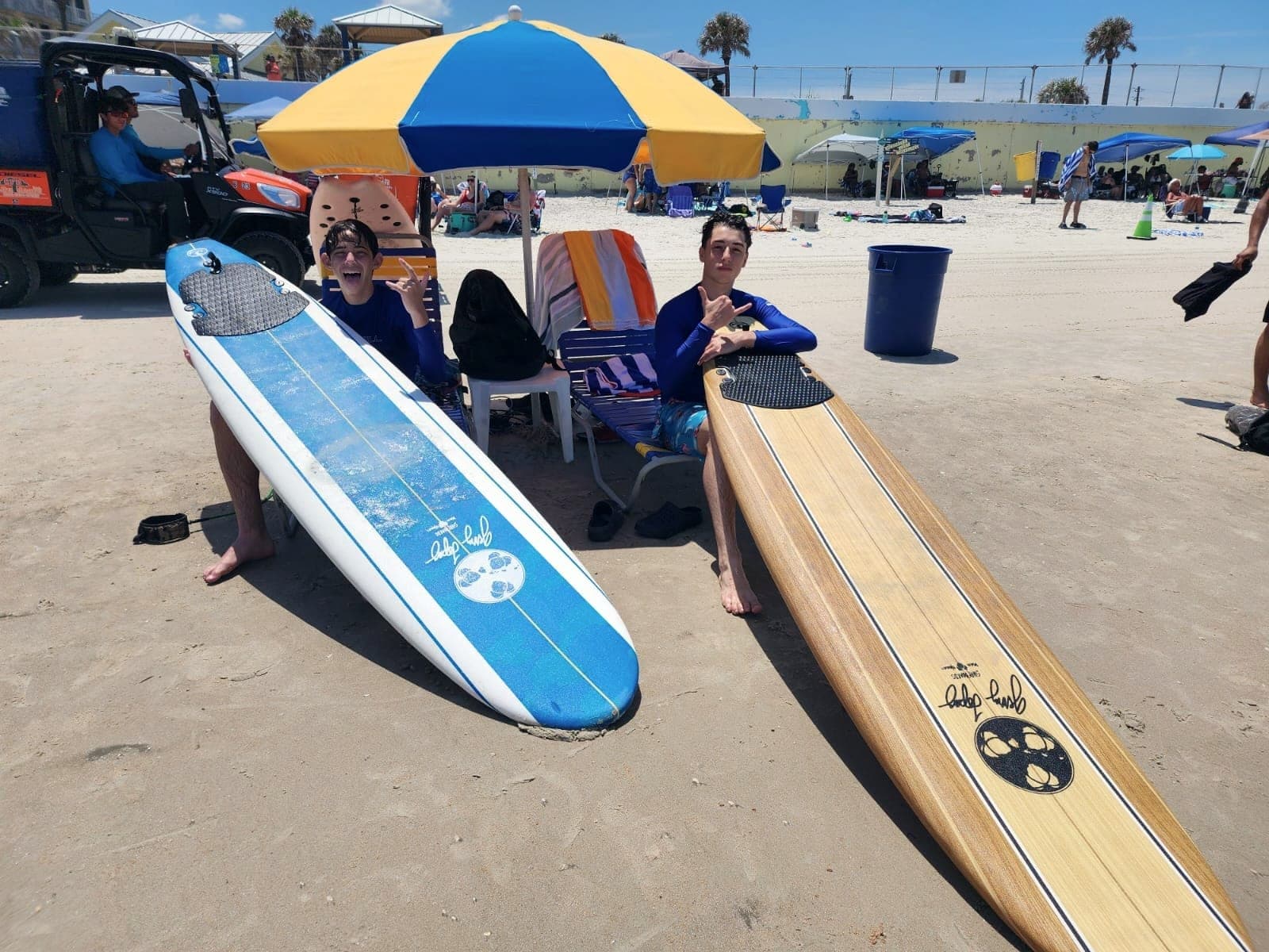 Two longboards standing in the sand beside a beach umbrella, with people sitting in the shade behind