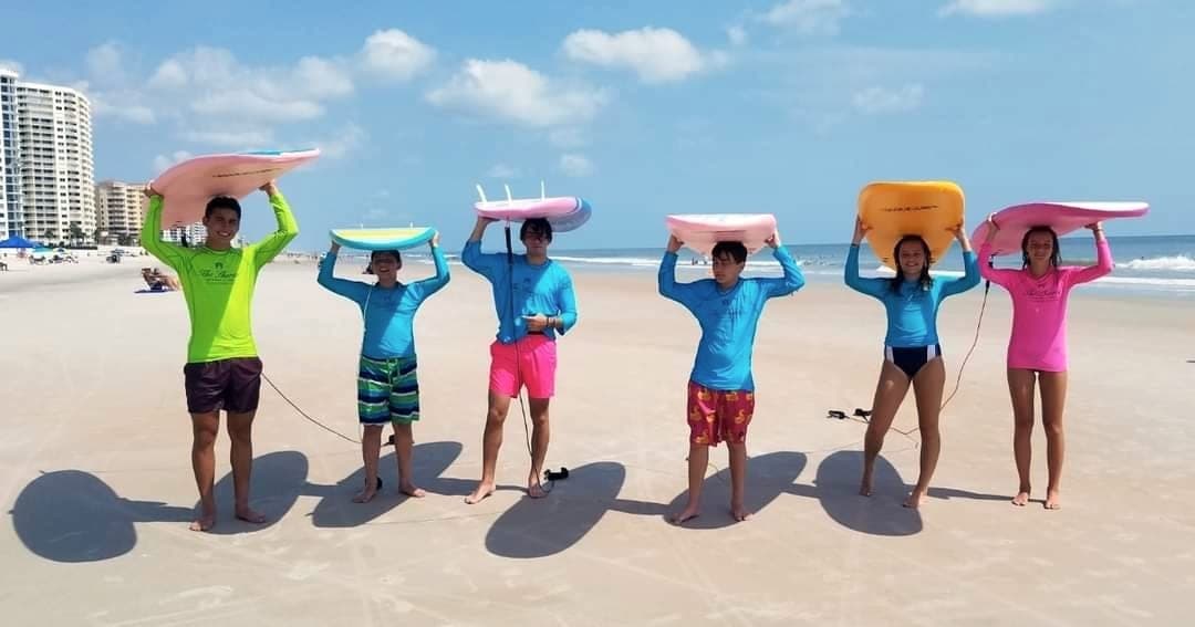 Students standing in a line on the sand with soft-top boards balanced on their heads