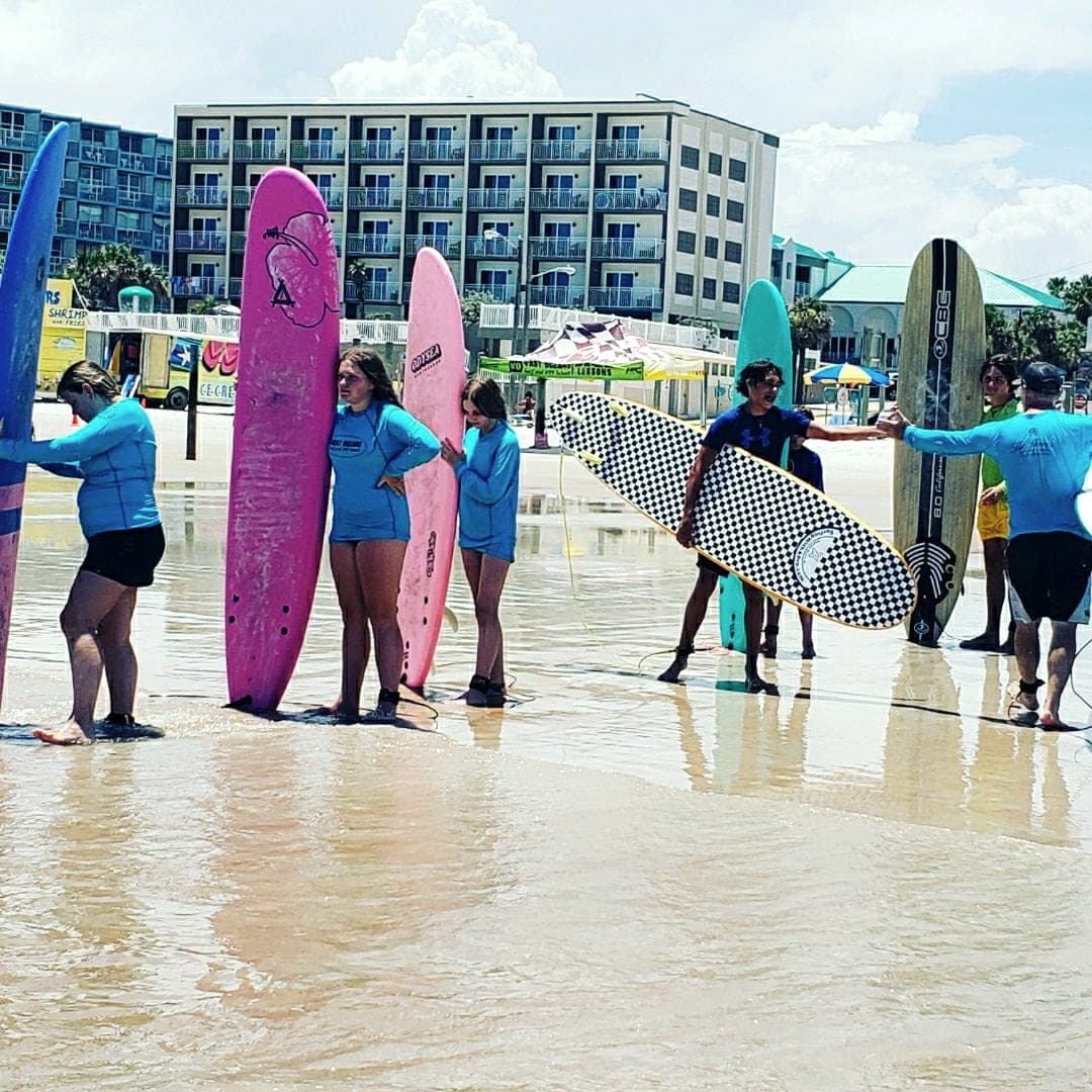 A lesson group with their boards at the waterline, hotels along the beach behind
