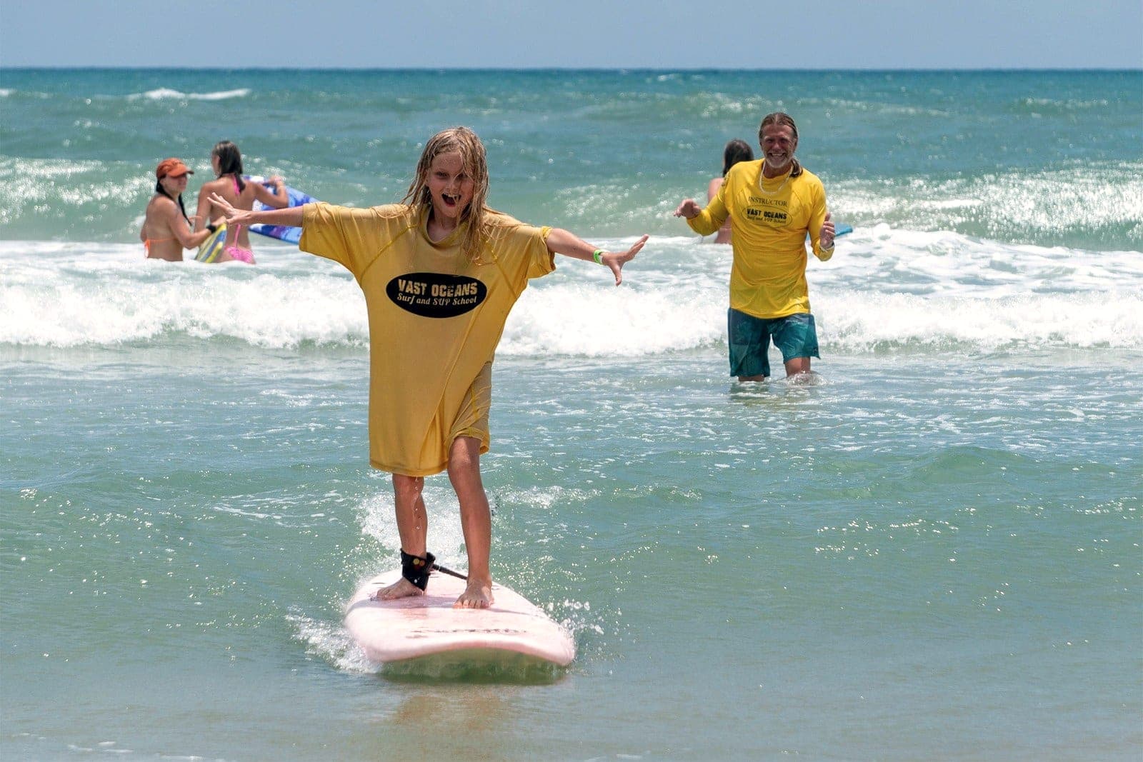 A young student standing on a board in shallow water with an instructor watching from behind