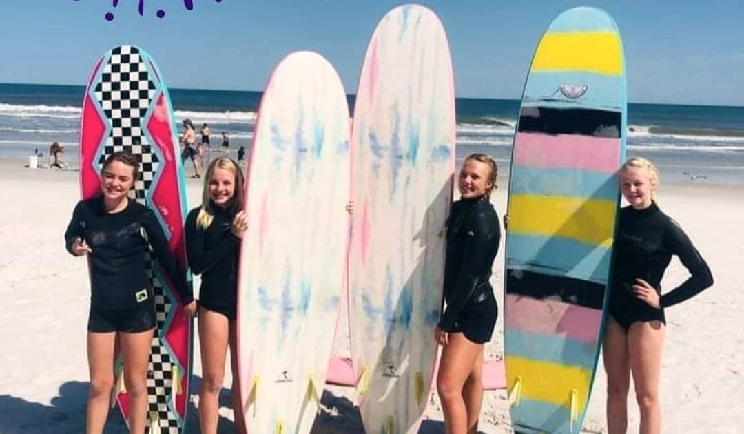 Five students standing in front of a row of upright soft-top boards on the beach