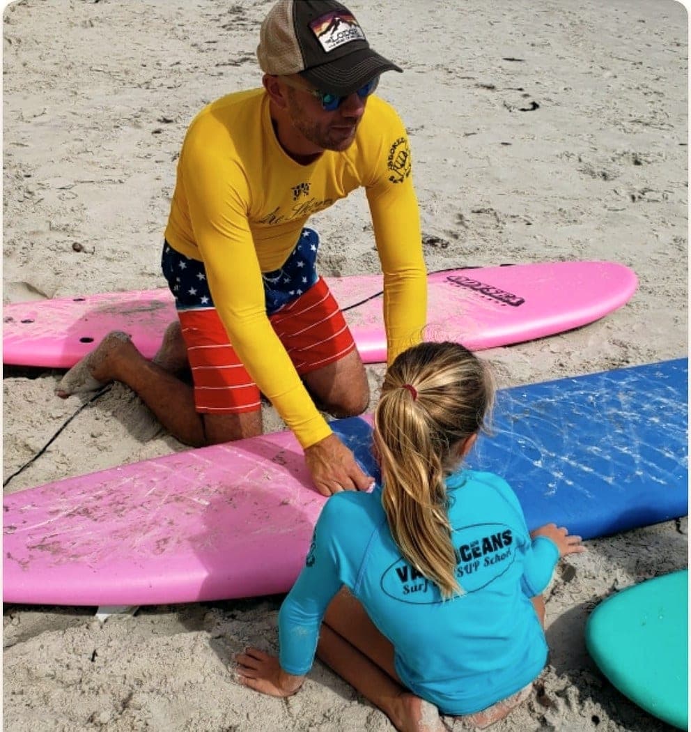 An instructor kneeling beside a young student on the sand between two soft-top boards