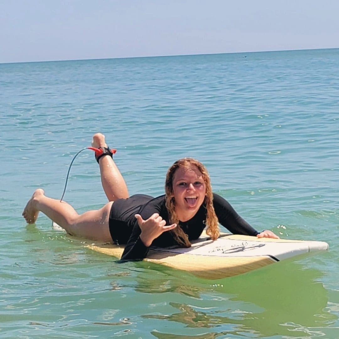 A student lying on a longboard in shallow water, throwing a shaka