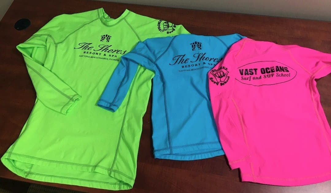 Three long-sleeve rash guards laid out on a table, one of them a Vast Oceans school shirt
