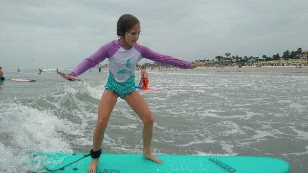 A young student standing on a soft-top board riding whitewater toward the beach