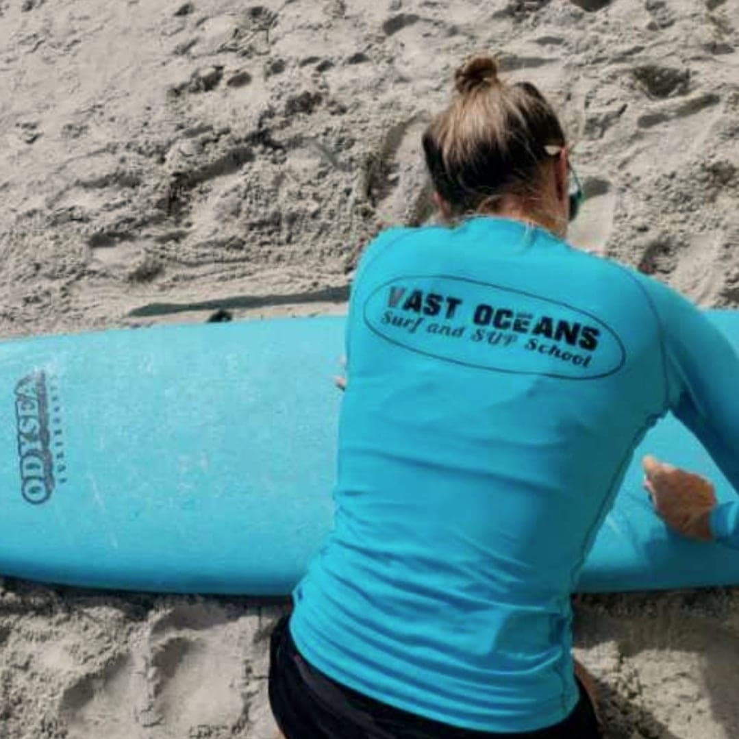 An instructor in a Vast Oceans shirt crouched over a board on the sand, seen from behind