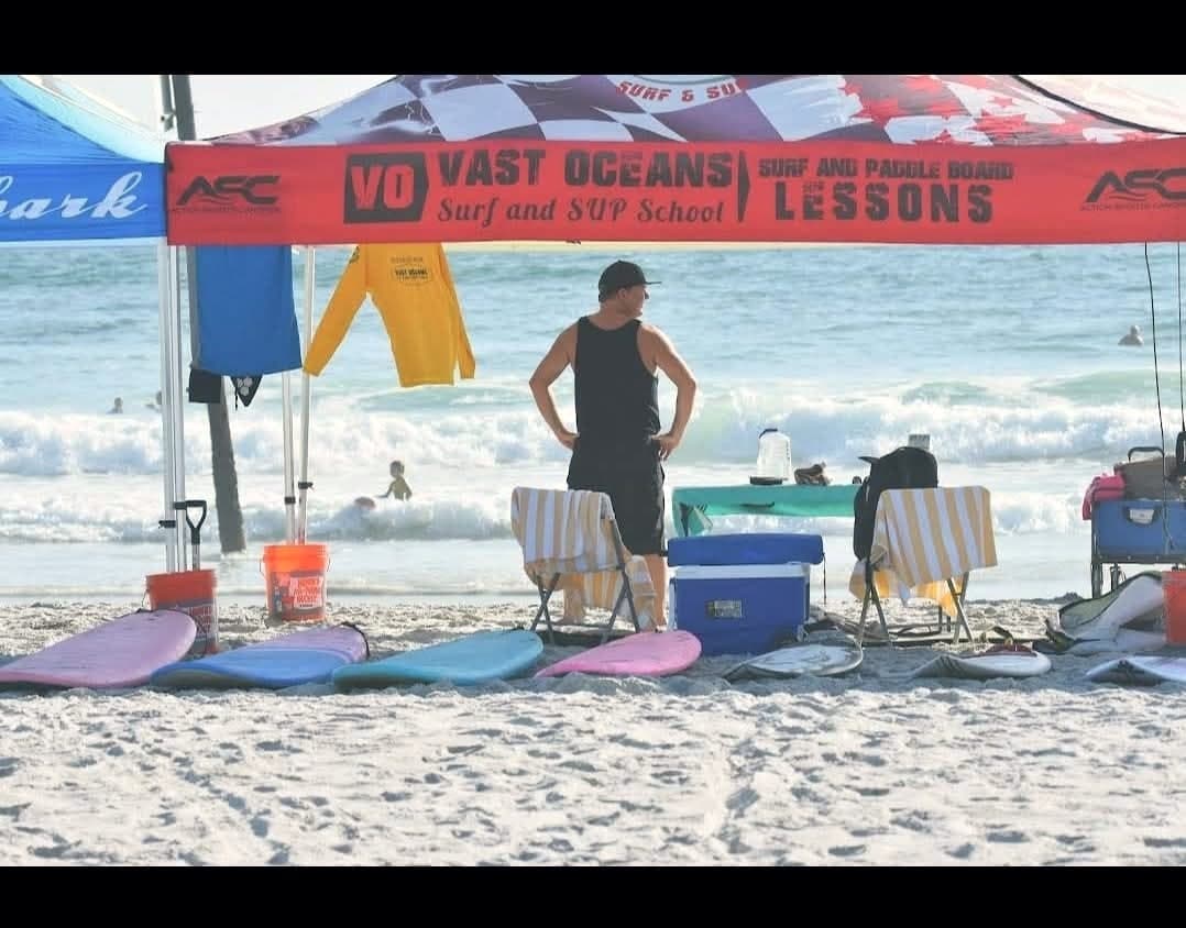 The Vast Oceans lesson tent with rash guards hanging and boards laid out on the sand, ocean behind