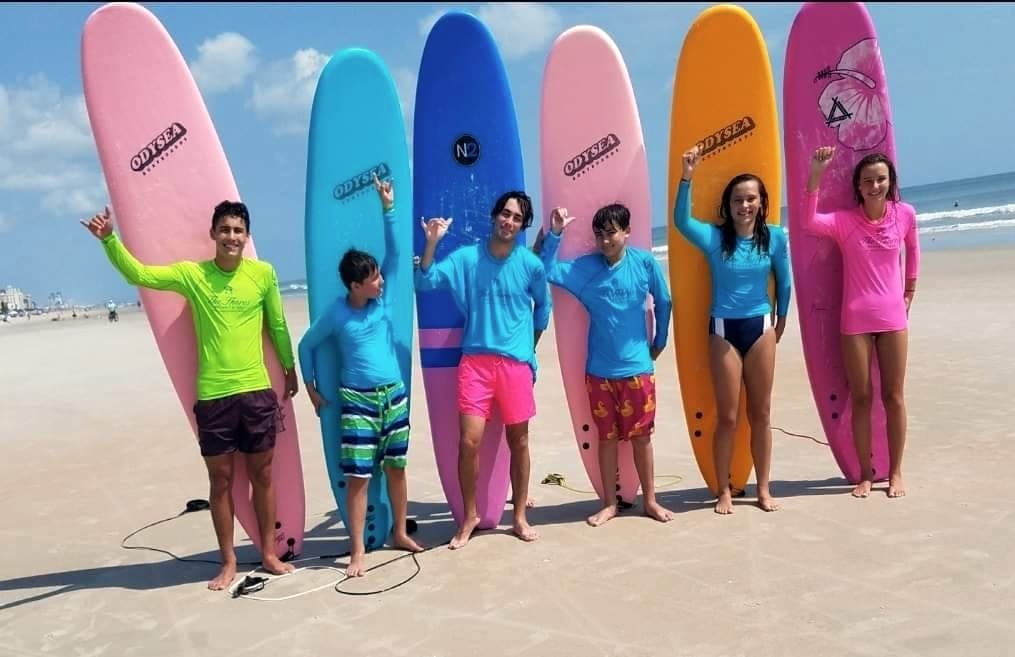 A class standing in front of a row of upright surfboards on the sand