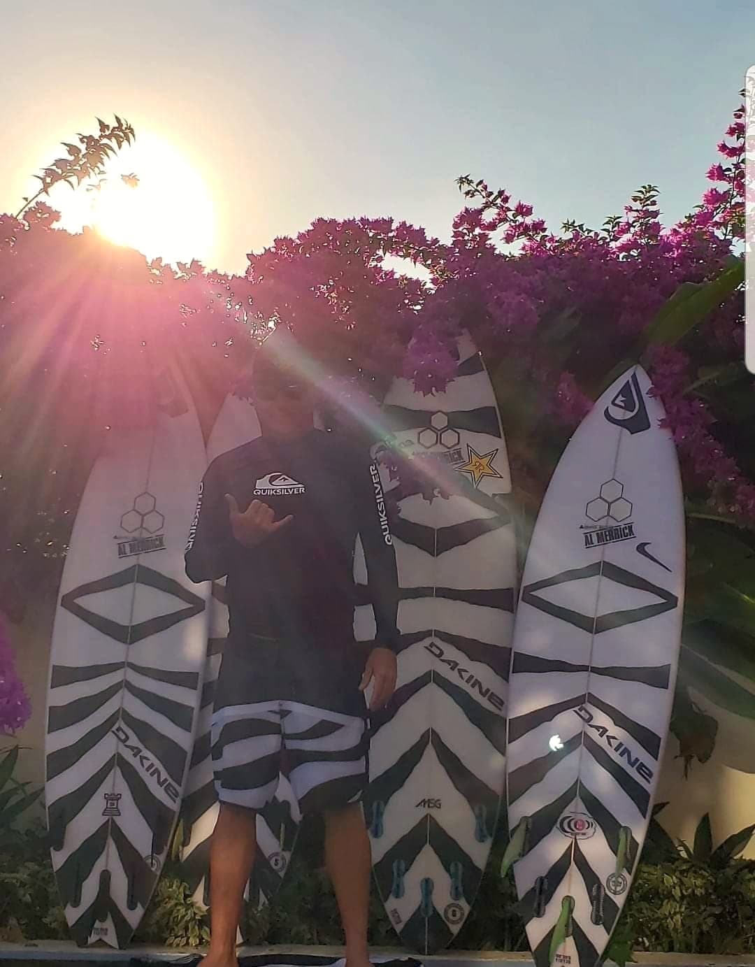 Ryan Ragan standing among four shortboards with the sun low behind bougainvillea