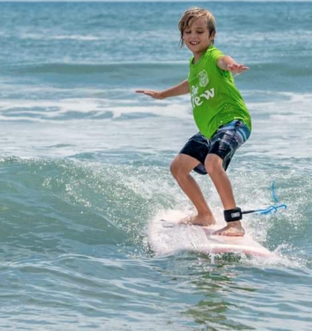 A young student riding a small wave on a soft-top board with both arms out for balance