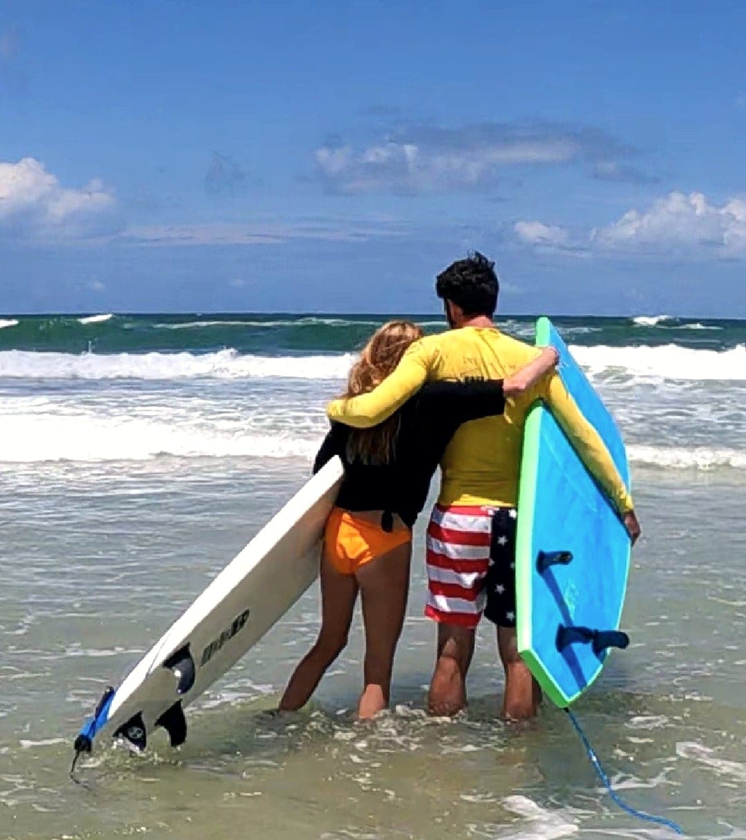 Two people seen from behind standing in shallow water holding surfboards